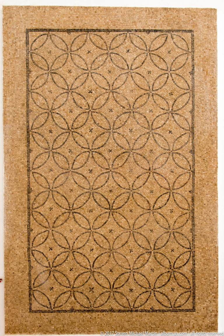 Rectangular Roman mosaic filled with repeated interlocking circles in dark tesserae on a warm ochre ground.