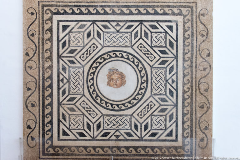 Black-and-white Roman mosaic with complex geometric borders surrounding a small central human face medallion.
