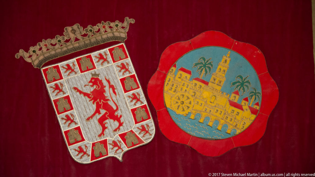 Two colorful heraldic emblems on red fabric, including a crowned lion shield and a fortified-city device.