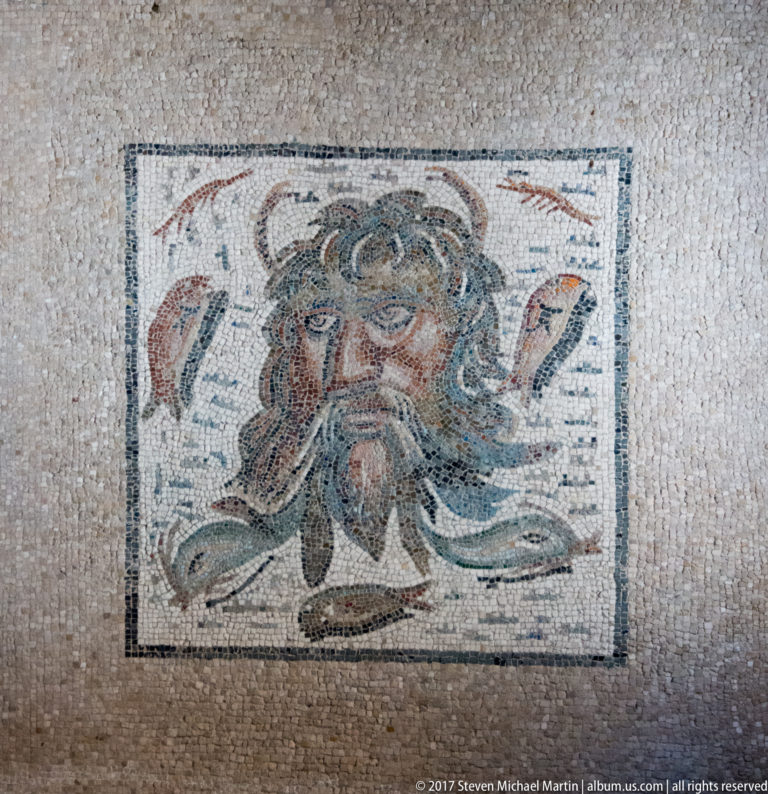 Roman mosaic panel of a bearded male face with curling hair and flowing beard inside a rectangular border.