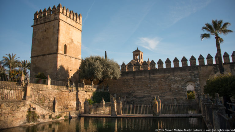 Square Alcázar tower and crenellated wall beside a narrow garden pool, framed by palms and clipped greenery.