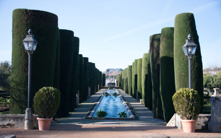 Straight water channel flanked by tall clipped cypress walls, lanterns, and potted shrubs in the Alcázar gardens.