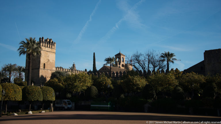 Sunlit square tower and crenellated Alcázar wall rise above palms, citrus trees, and clipped shrubs under a blue sky.