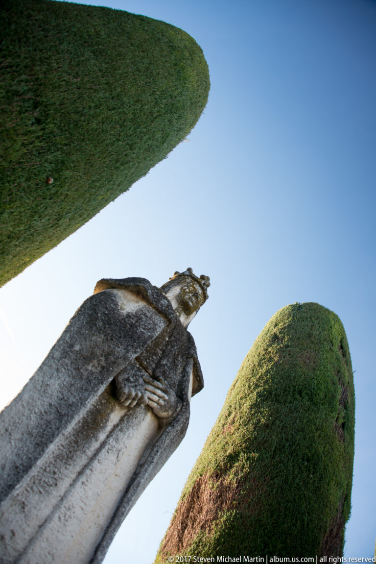 Low-angle close view of a weathered stone figure from the Columbus monument, framed between clipped cypress hedges.