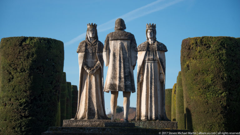 Stone statues of crowned Isabella and Ferdinand face the viewer while Columbus stands between them with his back turned.