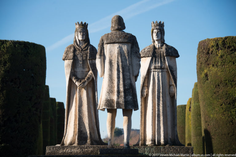 Centered stone group of Columbus between crowned Isabella and Ferdinand, enclosed by tall clipped cypress walls.