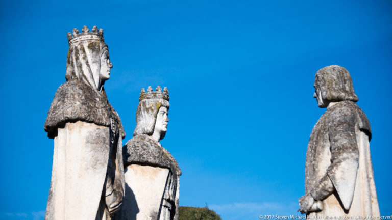 Side view of the stone Columbus monument with two crowned monarchs facing Columbus against a deep blue sky.