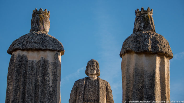 Rear view of the two crowned monarch statues with the front of Columbus visible between them against blue sky.