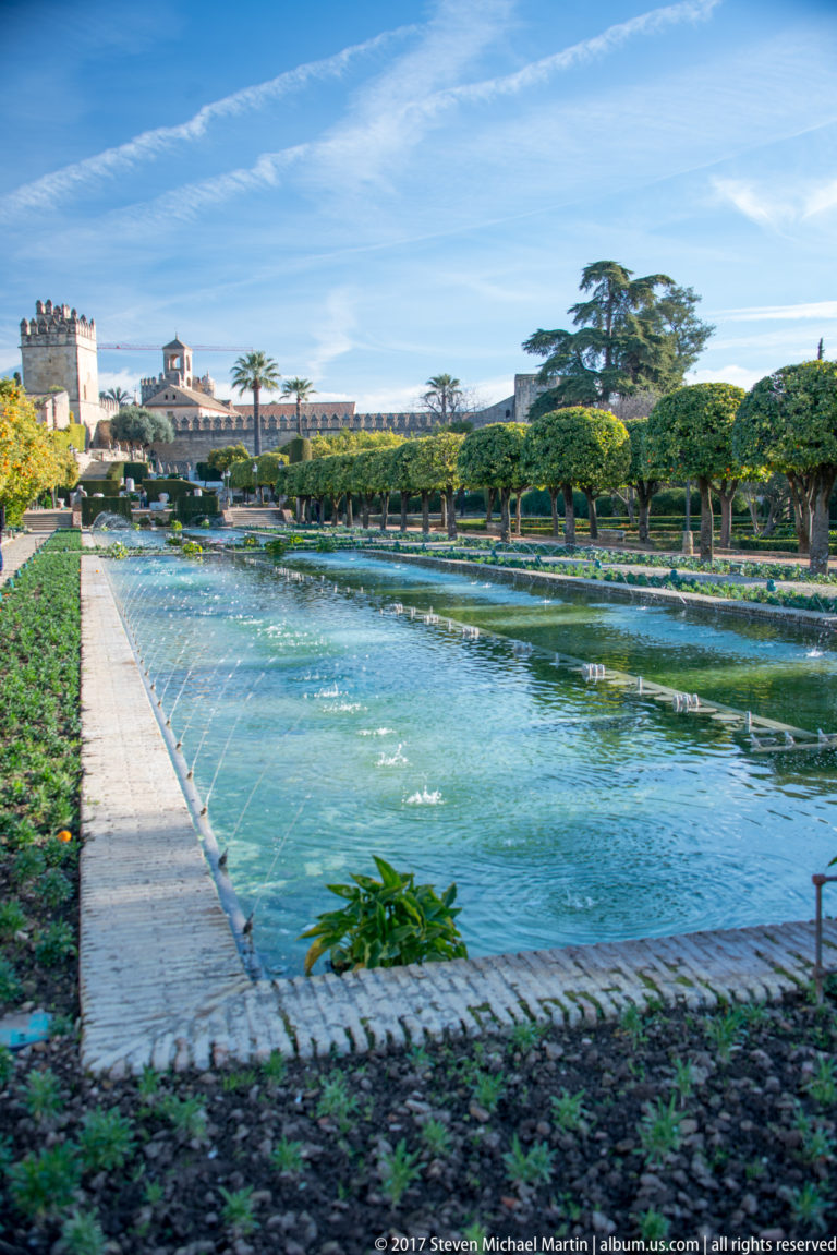 Broad rectangular pool with fountain jets, flower beds, clipped trees, and an Alcázar tower in the distance.