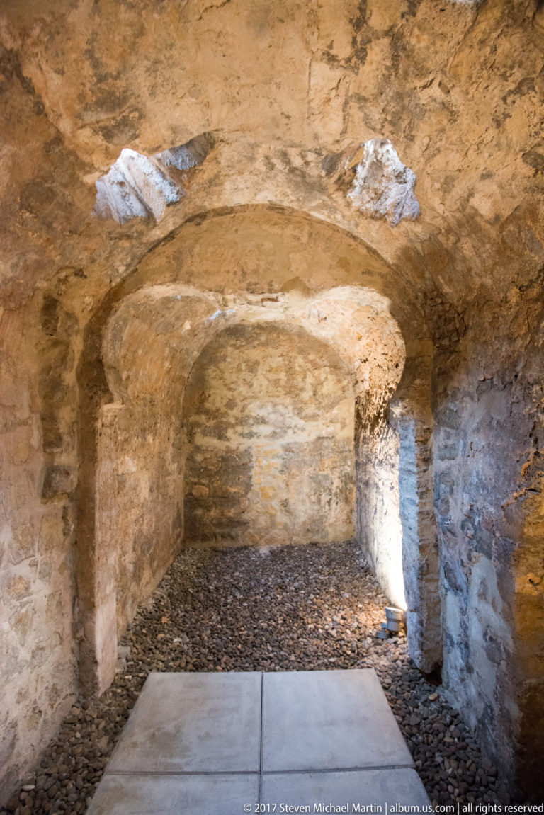 Rough excavated stone chamber with a low vault, rubble-covered floor, modern walkway, and discreet archaeological lighting.