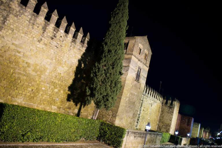 Floodlit crenellated stone wall and square tower of the Alcázar de los Reyes Cristianos beside a tall cypress at night.