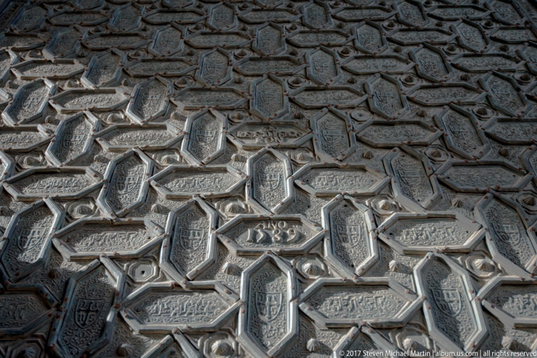 Close view of a dark carved wooden door covered with interlaced star patterns, polygons, and repeated small inscription panels.