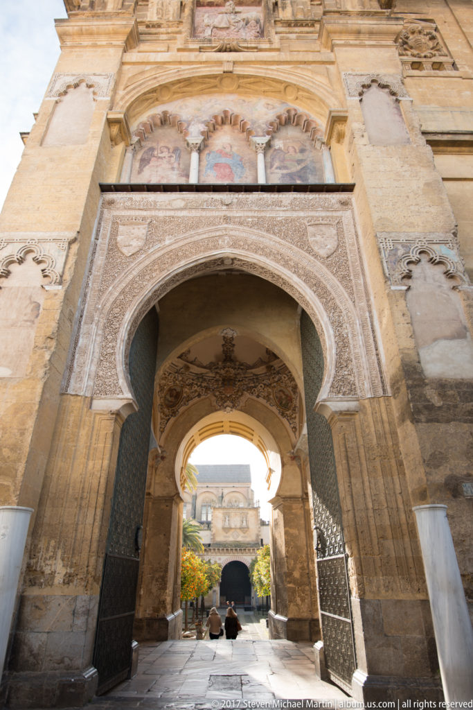 Large decorated horseshoe arch frames a second arch and sunlit courtyard, with carved geometric panels and stonework around the passage.