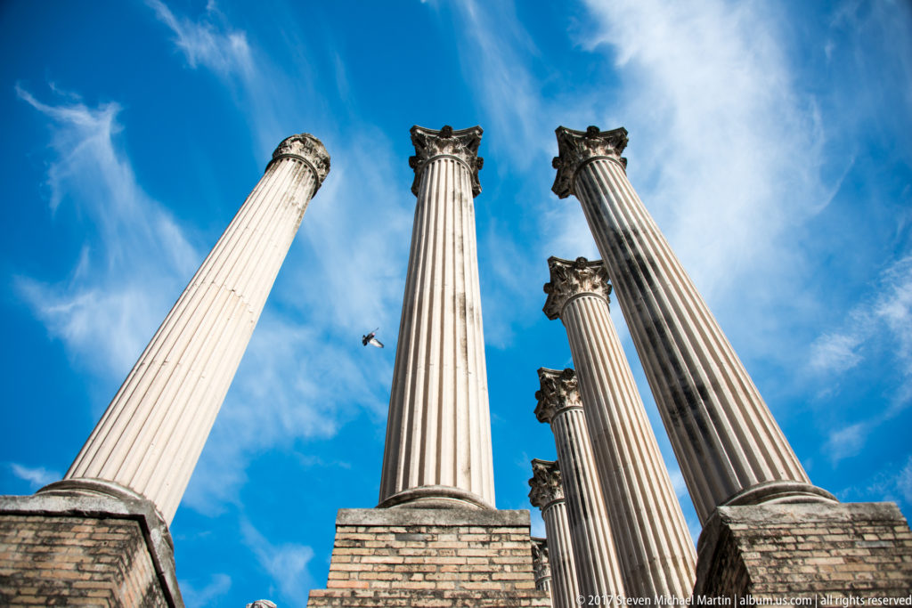 Extreme low-angle view looking up between fluted Corinthian columns of Córdoba’s Roman Temple into a bright blue sky.