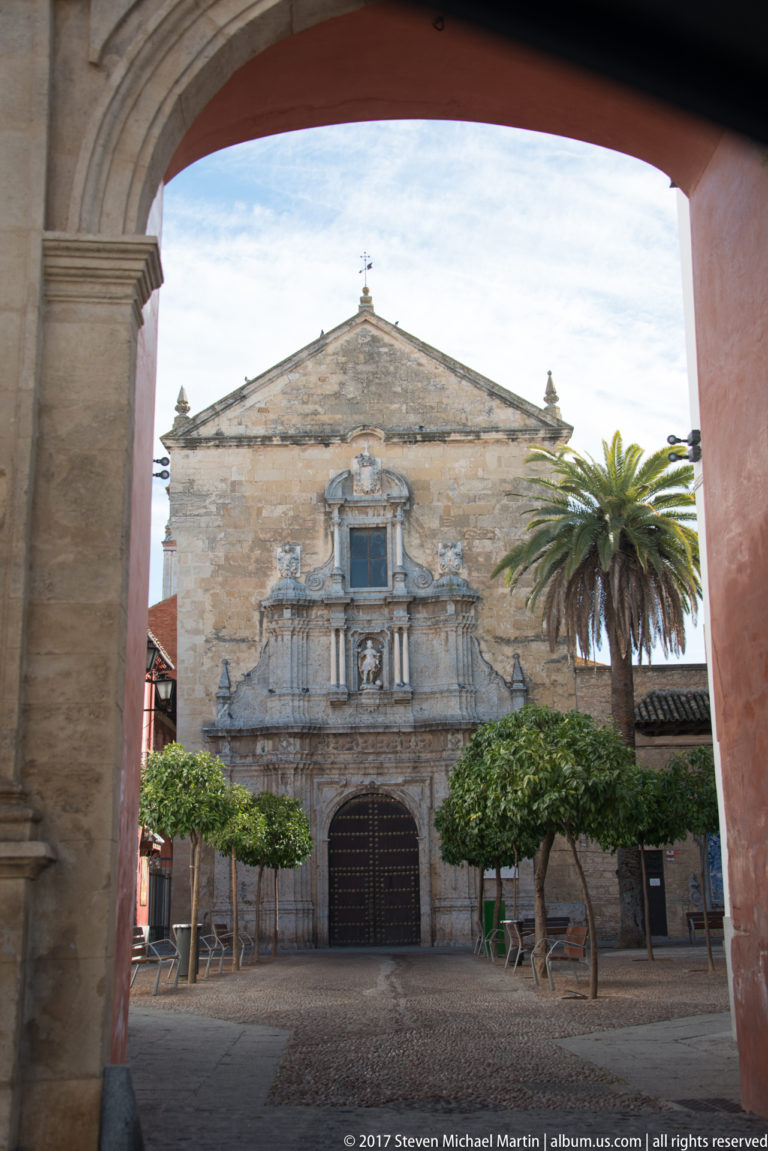 Stone church façade with elaborate Baroque portal, triangular gable, orange trees, and a tall palm, viewed through an archway.