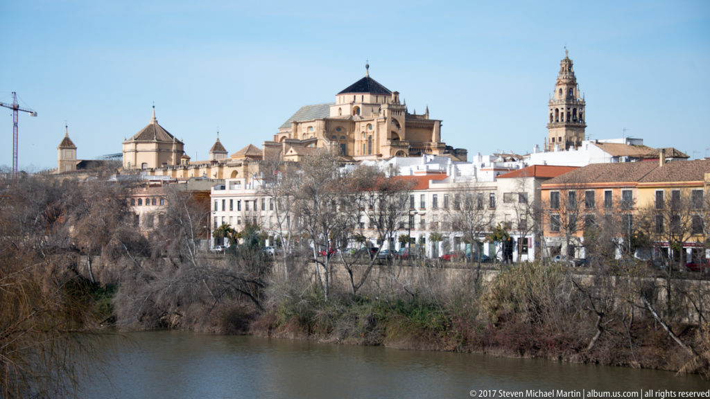Daytime skyline across the Guadalquivir with the Mosque-Cathedral’s cathedral roof, dome, and bell tower above Córdoba’s historic center.