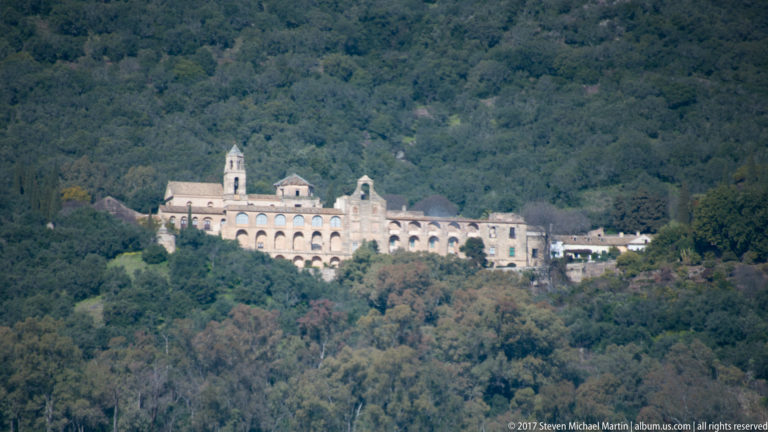 Long pale monastery complex with repeated arches, church front, and small bell tower set on a densely wooded hillside.