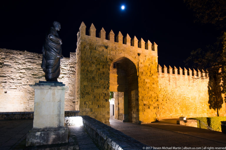 Floodlit crenellated stone gateway at the Alcázar, with a bronze statue on a pedestal in the foreground and moon above.
