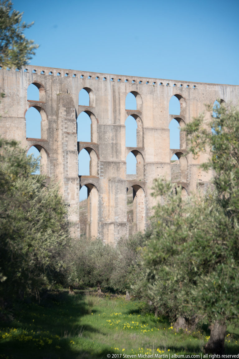Multi-tiered stone arches of the Amoreira Aqueduct rise behind olive trees and green ground under a clear blue sky.