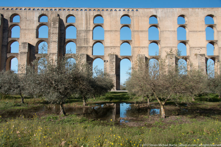 Frontal view of stacked Amoreira Aqueduct arches behind olive trees, with a small reflective water channel in the foreground.