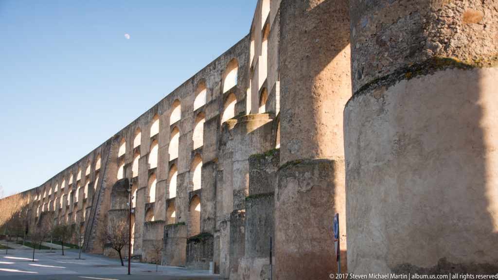 Street-level view along the Amoreira Aqueduct with repeated arches and massive projecting buttresses beside a paved walkway.