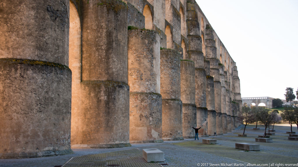 Close ground-level perspective along massive projecting buttresses supporting the Amoreira Aqueduct beside a paved plaza.