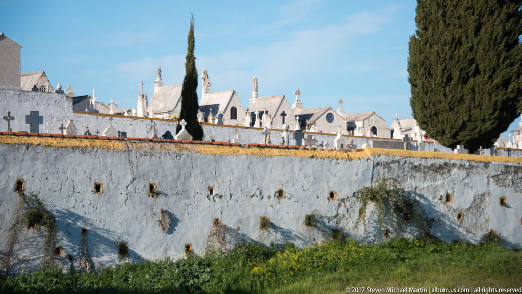 Whitewashed cemetery wall with pale tomb monuments and gabled structures beyond, flanked by tall dark cypress trees.