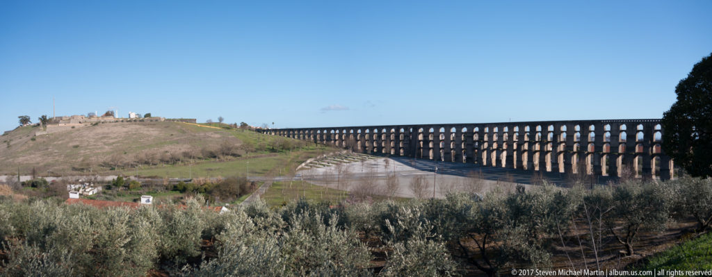 Wide landscape with olive trees, the long arcaded Amoreira Aqueduct, and fortified high ground beyond under a clear sky.
