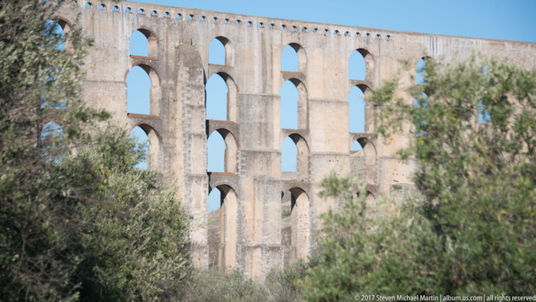 Close view of tall stacked arches and narrow piers of the Amoreira Aqueduct, partly screened by leafy trees.