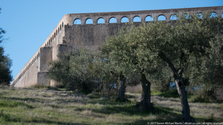 Oblique view along the long masonry Amoreira Aqueduct, with small upper arches and olive trees in the foreground.