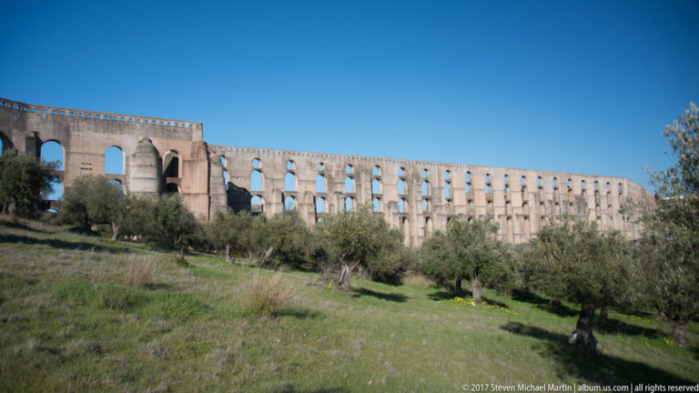 Curving stretch of the Amoreira Aqueduct above an olive grove, with a massive buttressed section at left.