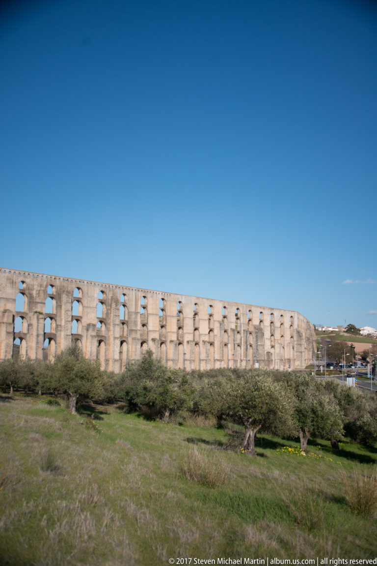 Wide panorama of the long multi-tiered Amoreira Aqueduct crossing open fields beneath a deep blue sky.