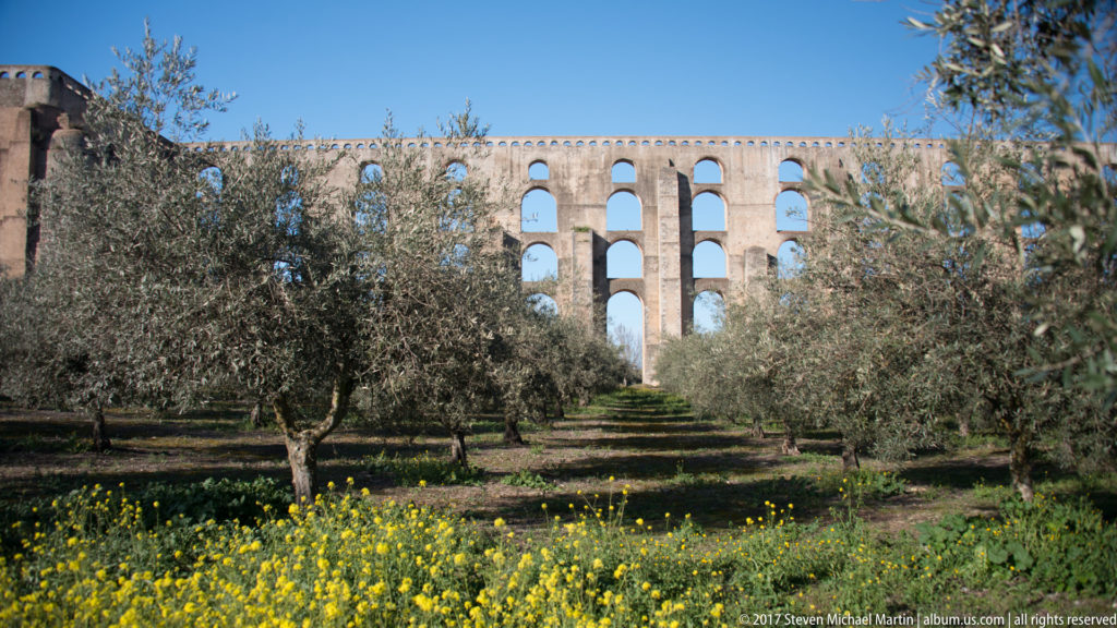 Olive trees and yellow wildflowers line a path toward the tall stacked arches of the Amoreira Aqueduct.