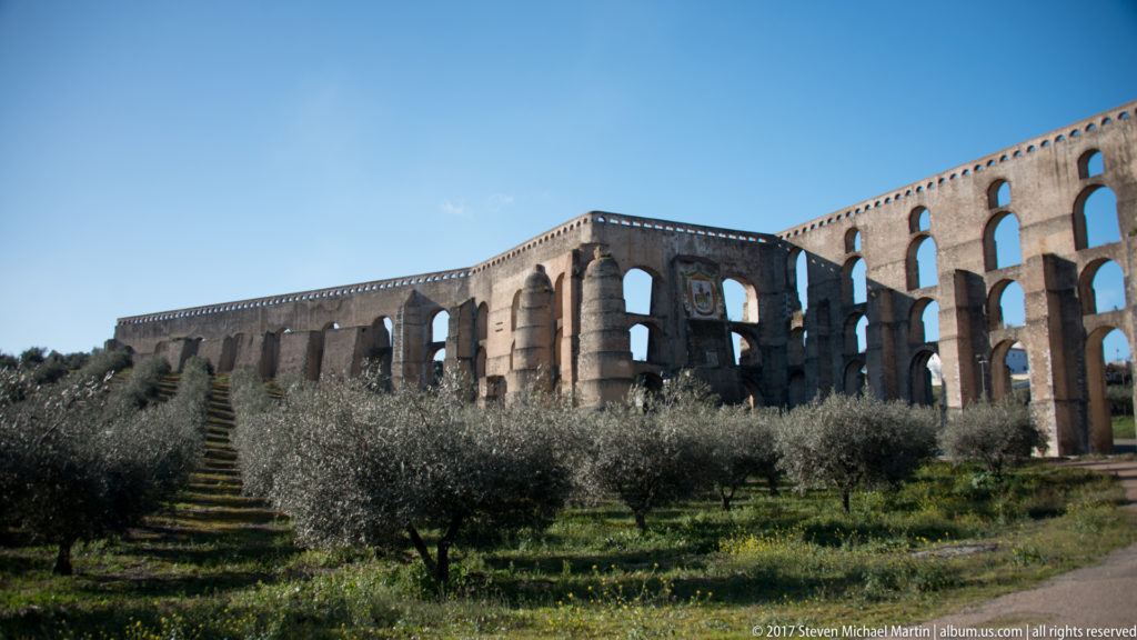 Two stretches of the Amoreira Aqueduct meet at a massive buttressed masonry section above olive trees.