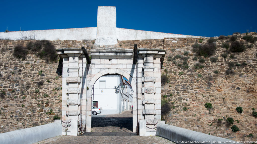 Round-arched stone gateway set in a rubble fortification wall, with a whitewashed street visible beyond.