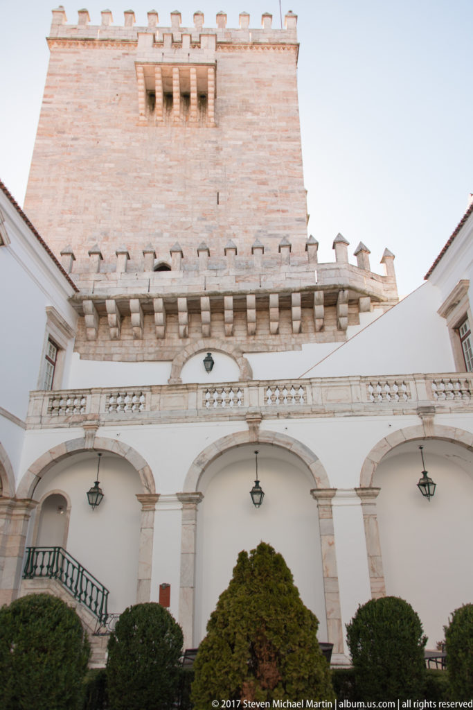 White arcaded courtyard with clipped shrubs and balustrade below a tall pale stone crenellated keep.