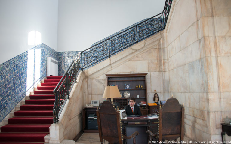 Marble staircase with red runner and dark iron railing rising beside a staffed reception desk and heavy wooden furniture.
