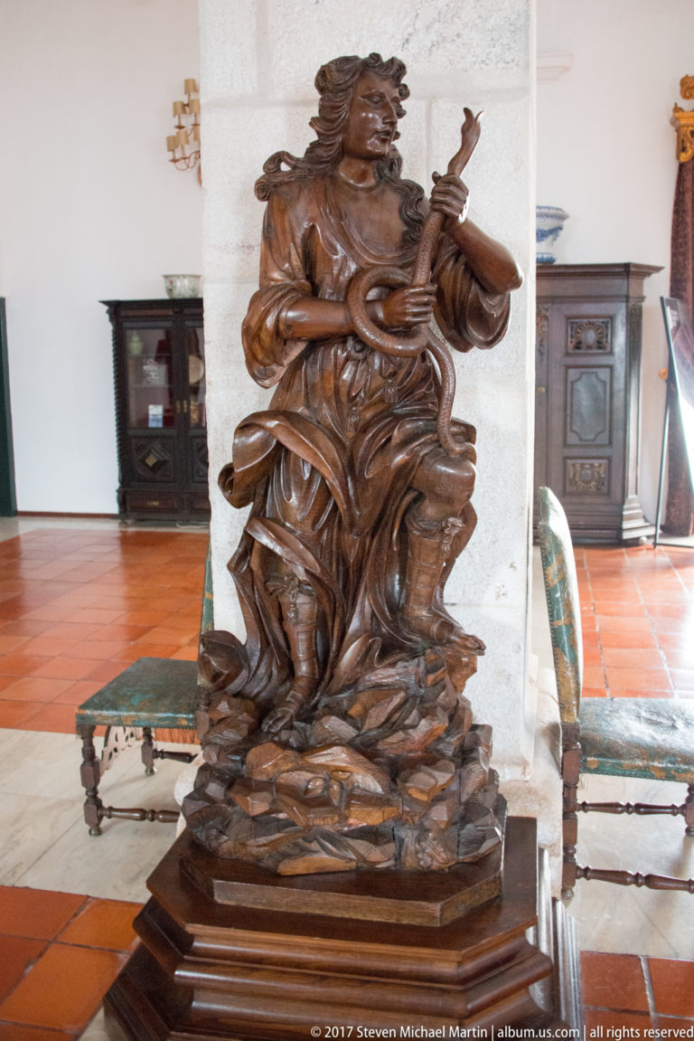 Carved wooden seated woman playing a small stringed instrument, displayed on a pedestal in a furnished historic interior.