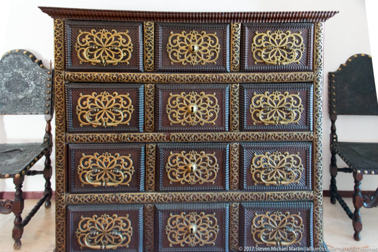 Large dark chest covered with repeating gold floral panels and patterned borders, flanked by historic chairs.