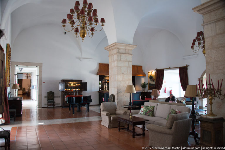 Vaulted white lounge with stone piers, sofas, chandeliers, terracotta floor tiles, and a black grand piano.