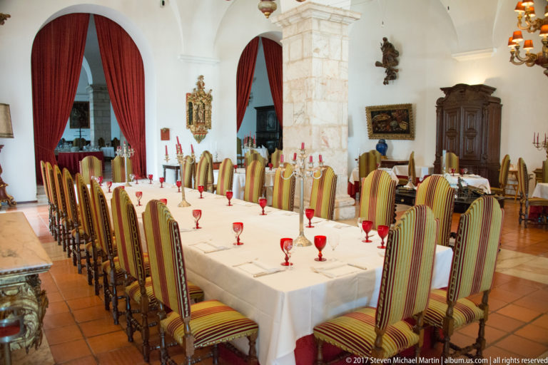 Formal dining room with long white-clothed tables, striped high-backed chairs, red goblets, stone piers, and red curtains.