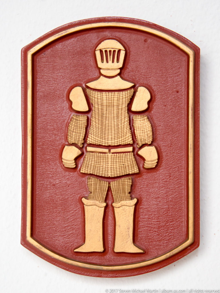 Cream-colored stylized armored figure in relief on a red shield-shaped plaque.