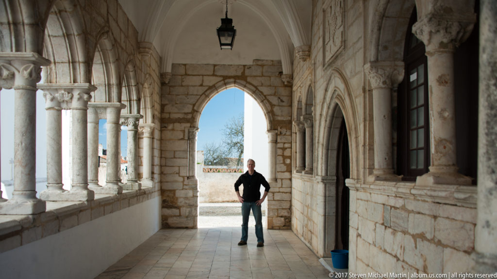 Stone arcaded gallery with slender columns, pointed arches, carved capitals, and a person standing in the sunlit end doorway.