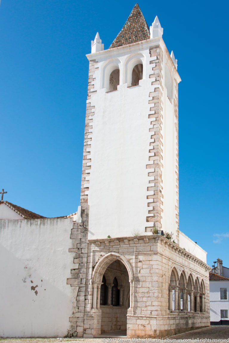 White square tower with stone corner blocks, arched openings at its base and top, and a pyramidal tiled roof.