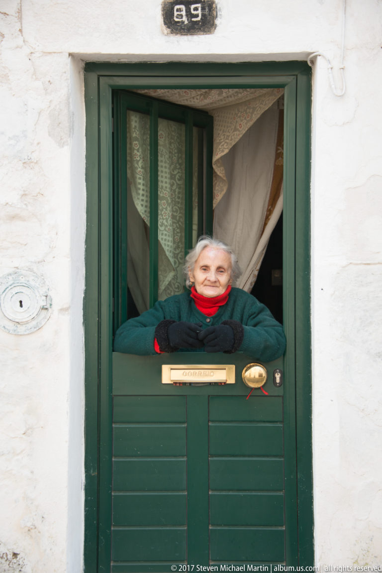 Elderly woman in a red scarf leaning over a dark green half-door in a whitewashed house numbered 89.