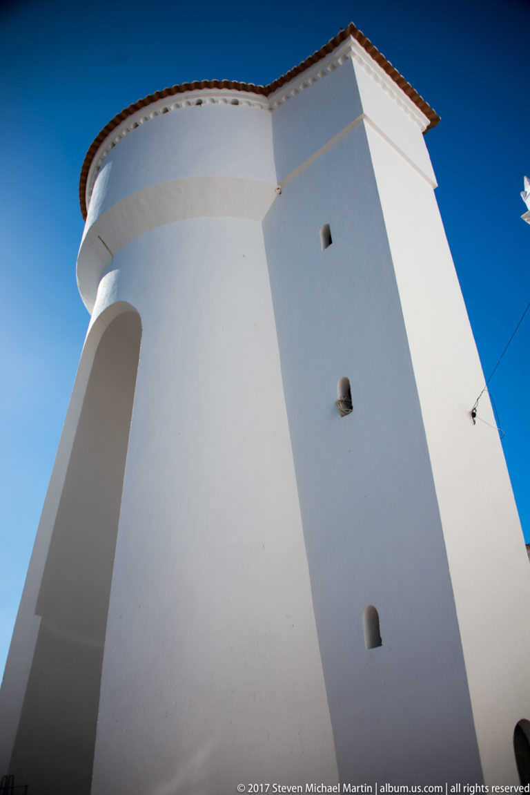 Low-angle view of a massive rounded white fortification tower with small openings and a red-tiled roof against blue sky.