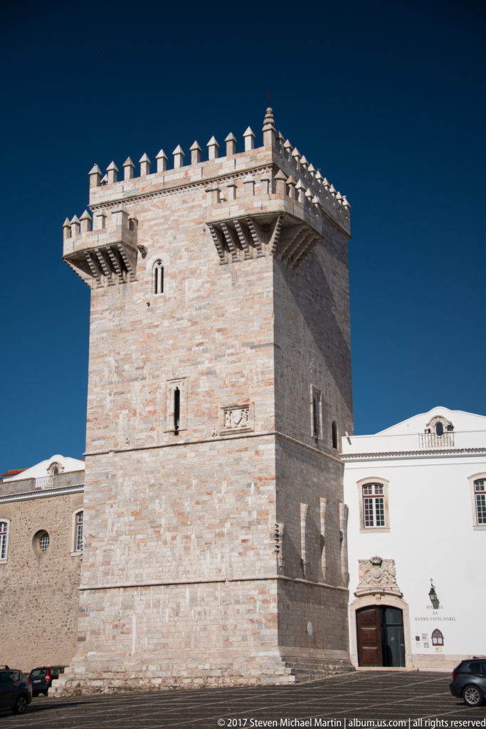 Square pale stone keep with narrow windows, deep corbelled crown, and pointed crenellations rising beside white palace buildings.
