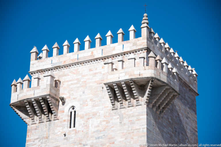 Close low-angle view of a pale stone keep’s corbelled parapet, pointed merlons, and narrow arched window.