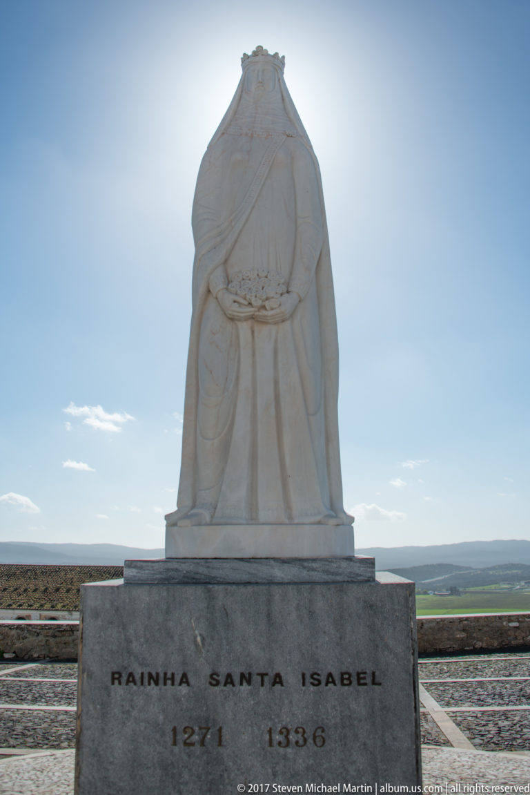 Backlit white statue of a veiled queen on a pedestal inscribed RAINHA SANTA ISABEL 1271–1336, overlooking open countryside.