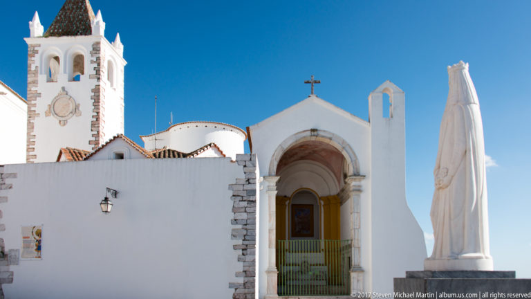 Whitewashed church with deep arched portal and green gate beside the tall white monument to Rainha Santa Isabel.